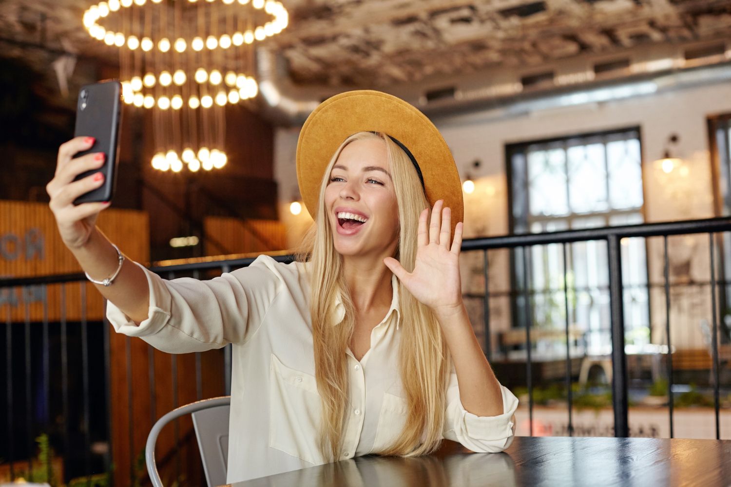 attractive young long haired blonde female sitting cafe table while lunch break making photo herself with her smartphone raising palm hello gesture smiling widely